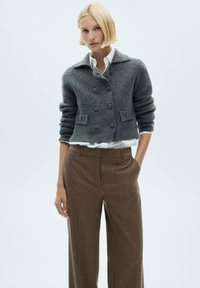 Šedý zkrácený cardigan s klopeným límcem, dvojitými knoflíky a strukturovaným pletením, spárovaný s hnědými kalhotami s kostkovaným vzorem.