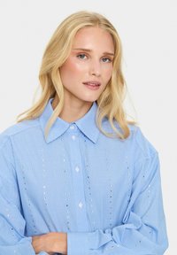 Camicia a righe blu e bianche con colletto classico, dotata di dettagli metallici decorativi lungo la parte anteriore e chiusure a bottone.