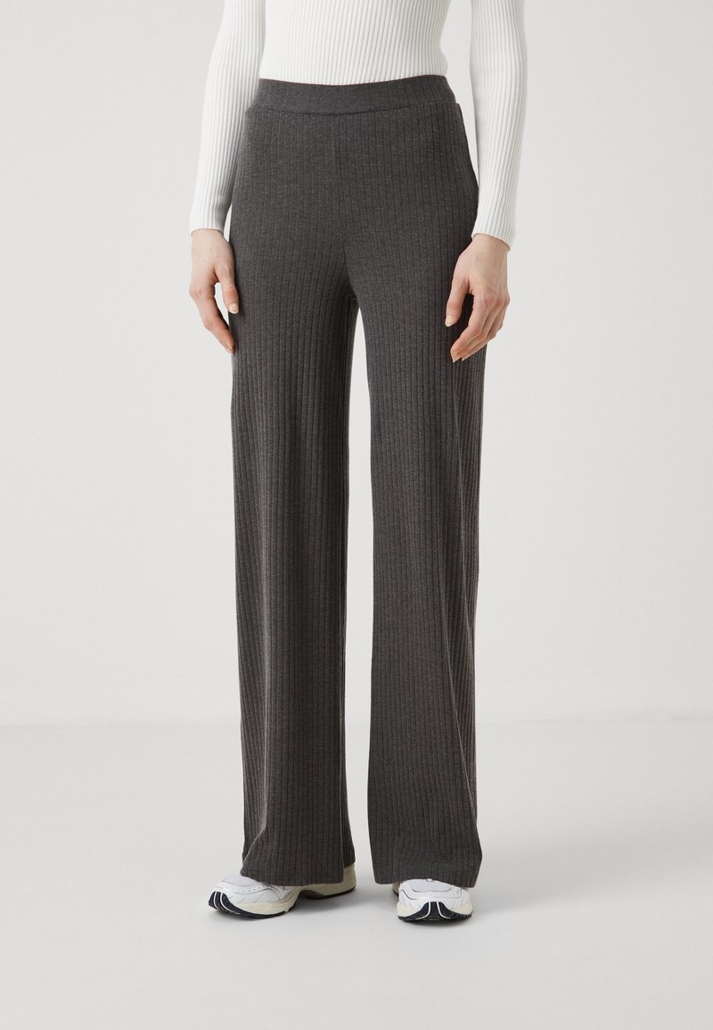 Femme portant un pantalon large côtelé gris foncé, un haut blanc à manches longues côtelé et des baskets blanches, debout devant un fond uni.