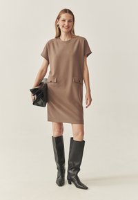 Braunes Kurzarmkleid mit zwei Taschen vorne und Knopf-Details. Das Model trägt kniehohe schwarze Lederstiefel und hält eine schwarze Clutch-Tasche.