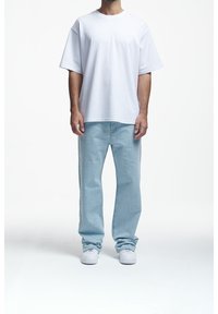 T-shirt bianca a maniche corte in cotone abbinata a jeans dritti azzurri e sneakers bianche, in piedi contro uno sfondo neutro.