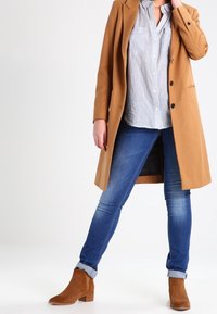 Camelullrock med strukturerade slag, grå randig button-up skjorta, blå denimjeans och bruna mocka ankelskor med klack.