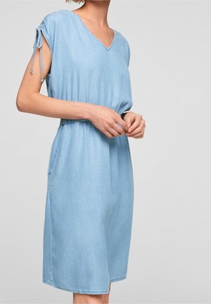 Vrouw draagt een lichtblauwe mouwloze jurk tot op de knie met een V-hals en een detail van geknoopte schouders, staand met de handen in elkaar gevouwen.