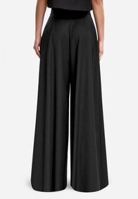 Czarne szerokie spodnie o wysokim stanie, wykonane z gładkiego materiału; mają fałdy dla zwiększenia objętości i swobody. Pokazany widok z tyłu.