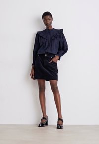 Marine gestreiftes Blusenshirt mit Rüschensaum, kombiniert mit einem schwarzen Minirock. Das Modell trägt schwarze High Heels und steht vor einem neutralen Hintergrund.