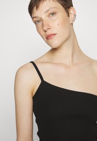 Top preto canelado sem mangas com alças finas, apresentando um decote assimétrico. Modelo exibe um design limpo e minimalista com uma textura suave.