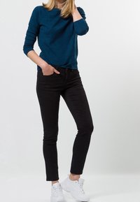 Turkooise top met lange mouwen en een textuurpatroon, gecombineerd met zwarte skinny jeans en witte sneakers, voor een casual en moderne outfit.