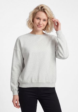 Kvinde med blondt hår iført en lysegrå sweatshirt og sorte bukser, smilende med den ene hånd, der rører ved hendes hår mod en hvid baggrund.