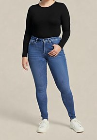 Personne portant un haut noir à manches longues, un jean skinny bleu et des baskets blanches, debout devant un fond beige uni.