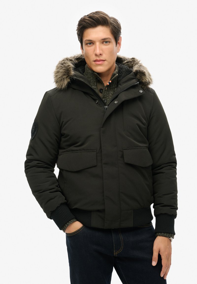 Veste isolée noire avec capuche doublée de fourrure, deux poches avant, poignets côtelés et texture de tissu lisse. Portée sur un pull en tricot.