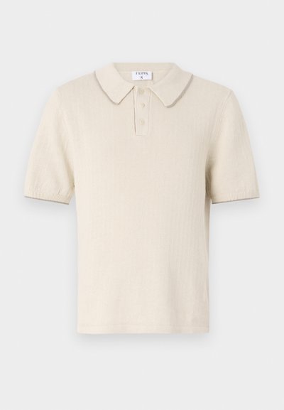 Polo de creme com mangas curtas, textura ribada, pala com três botões e colarinho em contraste. O material parece suave e leve.