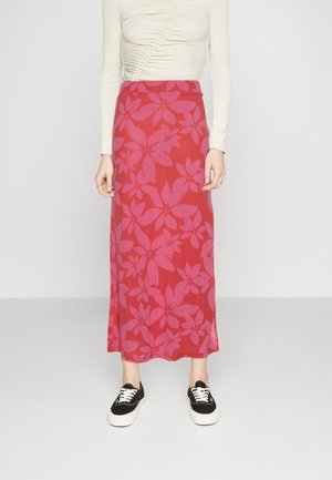 Vrouw draagt een rode maxirok met roze bloemenpatroon, een crèmekleurige longsleeve met plooitjes en zwarte sneakers met witte zolen en veters.