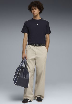 Młody mężczyzna w czarnej koszulce Puma, beżowe szerokie spodnie, czarny pasek, trzyma granatowo-białą torbę typu duffle, stoi na szarym tle.