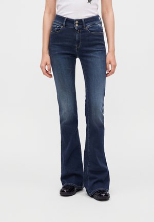 Person iført mørkeblå vidde jeans med to knapper og sorte sko, kombineret med en hvid top synlig fra taljen og ned.
