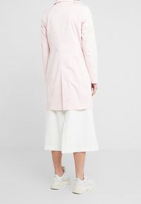 Henkilö, joka on pukeutunut vaaleanpunaiseen pitkään takkiin, valkoisiin leveälahkeisiin keskipitkiin housuihin ja valkoisiin paksupohjaisiin lenkkareihin, seisoo yksinkertaista vaaleaa taustaa vasten.