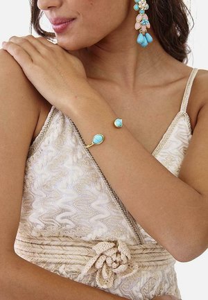 Mujer con top de encaje crema con una pulsera dorada con piedras turquesas y pendientes florales a juego en turquesa y oro.