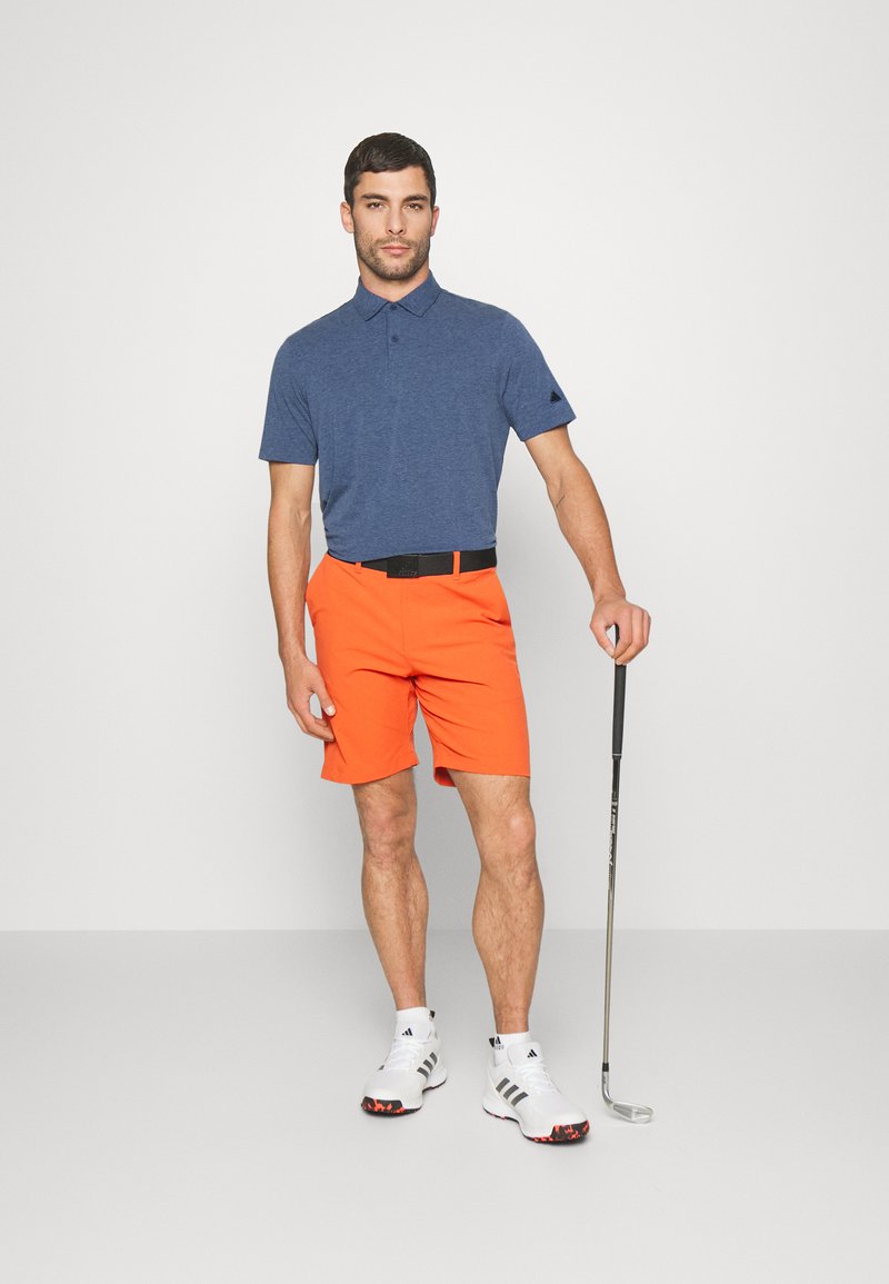 Hombre vistiendo un polo azul, pantalones cortos naranjas, zapatos de golf blancos, sosteniendo un palo de golf, de pie contra un fondo claro y liso.