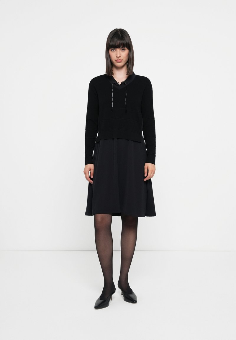 Schwarzer Pullover mit Kapuzen-Design, kombiniert mit einem fließenden schwarzen Kleid; beide Teile weisen eine glatte Textur und eine taillierte Silhouette auf.