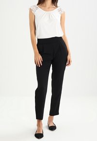 Kvinna i vit ärmlös topp med spetsaxlar, svarta högmidjade smalna byxor och svarta spetsiga ballerinas, stående mot en enfärgad bakgrund.