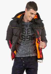 Schwarze Daunenjacke mit orangefarbiger Innenseite und roten Akzenten, ausgestattet mit einem Reißverschluss und elastischen Bündchen. Model trägt ein T-Shirt mit Grafik darunter.