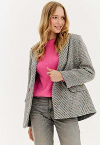 Texturovaný, vícebarevný blazer vyrobený z tkaniny s vzorem rybí kosti; má růžové knoflíky a je spárován s růžovým svetrem.