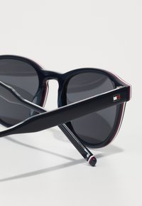 Ochelari de soare navy cu lentile închise, cadru rotund și brațe negre elegante, având un logo mic și un accent roșu-alb aproape de vârfuri.
