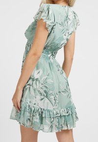 Robe fleurie vert clair avec des manches courtes volantées, une taille élastiquée et un ourlet à volants superposés. Fabriquée en tissu léger avec une texture lisse.