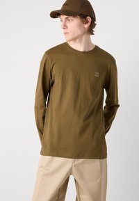 Langärmliges olivgrünes T-Shirt mit Rundhalsausschnitt und dezentem Logopatch. Weicher Baumwollstoff, kombiniert mit beigen Hosen und einer passenden Kappe.