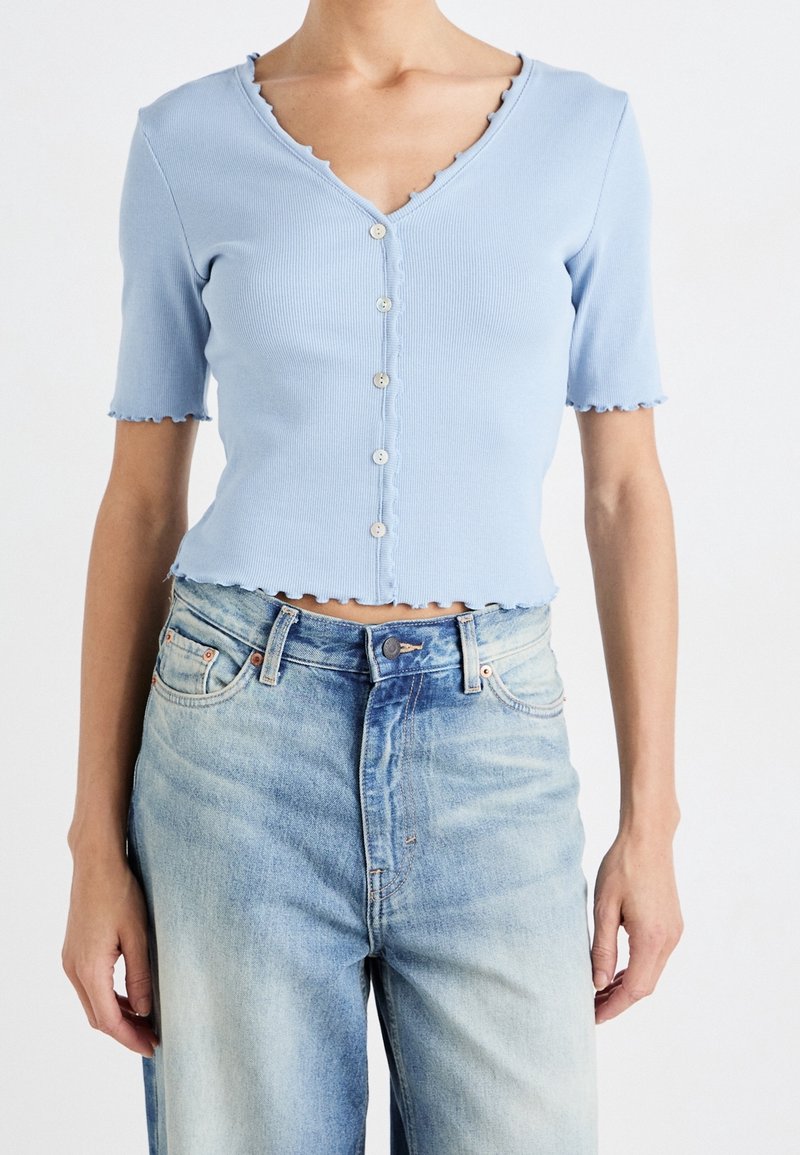 Femme portant un cardigan court bleu clair boutonné, manches courtes, et un jean bleu clair taille haute sur fond blanc.