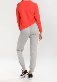 Rotes Langarmhemd, graue Jogginghosen mit elastischen Bündchen und schwarze Sportschuhe. Das Hemd hat einen leicht höheren hinteren Saum.
