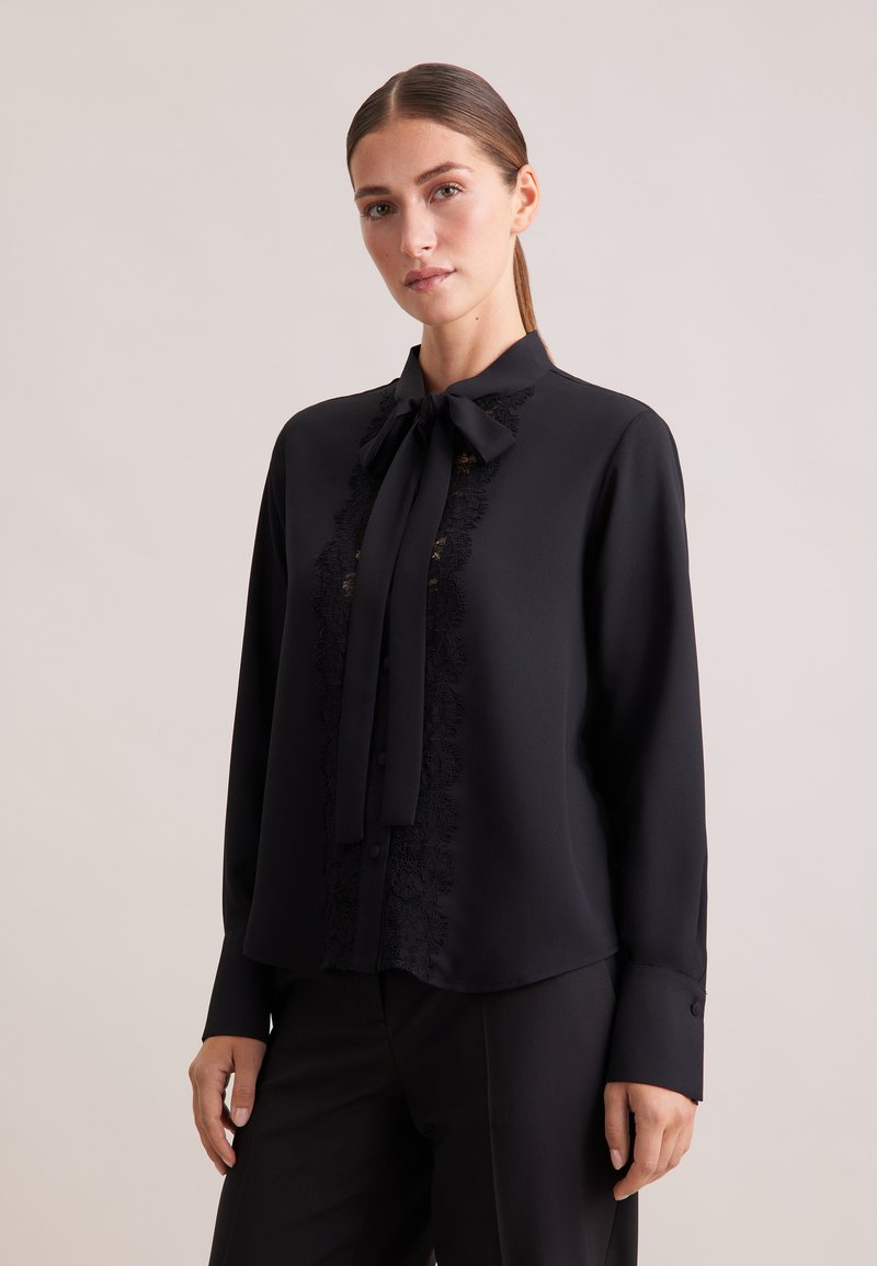 Blusa nera con fiocco al colletto, caratterizzata da dettagli in pizzo intricati lungo la parte frontale, maniche lunghe e polsini con bottoni. Tessuto liscio.