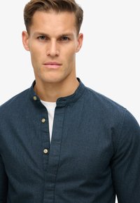 Donkerblauwe overhemdd met een Mandarin-kraag, textuurstof en twee knopen bij de halslijn. Gelaagd over een wit shirt.
