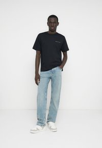 Μαύρο βαμβακερό μπλουζάκι με κοντά μανίκια και μικρό εκτυπωμένο λογότυπο στο στήθος. Ανοιχτό μπλε τζιν τζιν με ίσια γραμμή. Άσπρα αθλητικά παπούτσια.