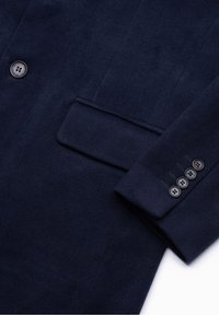 Manche d’un manteau en laine bleu marine avec quatre boutons noirs et une poche à rabat à l’avant, montrant la texture et les détails de couture.