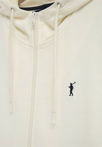 Cream-colored zip-up hoodie with drawstrings and a small black embroidered golfer logo on the chest.