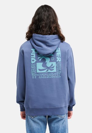 Personne aux longs cheveux bouclés portant un sweat à capuche bleu avec un motif abstrait de palmier bleu clair et un texte dans le dos, associée à un jean bleu.