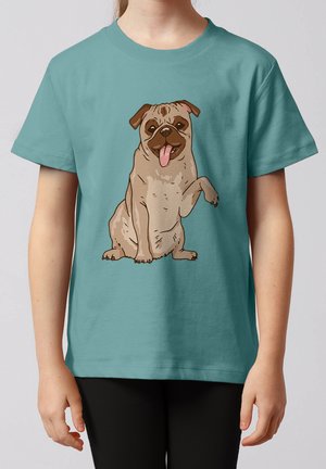 Kind trägt ein türkisfarbenes T-Shirt mit einem aufgedruckten, sitzenden Cartoon-Mops mit herausgestreckter Zunge und einer erhobenen Pfote auf der Vorderseite.
