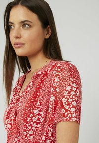 Blouse rouge à fleurs avec manches courtes, un col et un tissu léger et texturé orné de motifs floraux blancs.