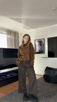 Nainen ruskeassa neuleessa ja tummissa leveälahkeisissa housuissa seisoo olohuoneessa, jossa on televisio, kehystetty taide, musta säkkituoli ja harmaa matto.