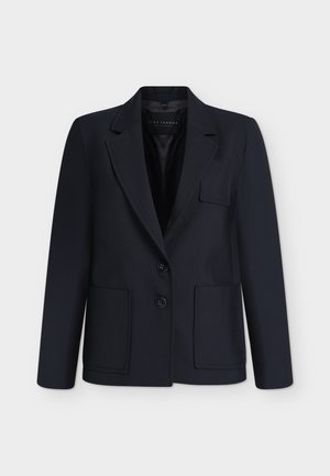 Personne portant un blazer noir, une chemise blanche, un pantalon beige et des talons pointus noirs tenant un sac à main beige sur un fond blanc uni.