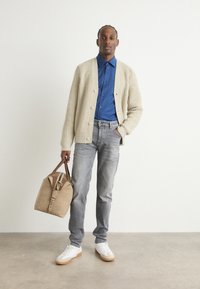 Cárdigan de canalé beige sobre una camisa azul de botones, jeans grises y zapatillas blancas. En una mano se sostiene un bolso marrón y beige con asas.