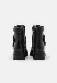 Une paire de bottines en cuir noir avec des semelles épaisses à profil cranté et des détails de poche sur le côté, vues de l'arrière sur un fond blanc.