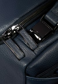 Donkerblauw leren tasdetail met dubbele ritsen voorzien van zilverkleurige hardware en een gestructureerde afwerking. Opvallende merknaam op de ritstrekkers.