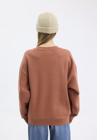 Kovově červený oversized svetr s dlouhými rukávy, vyrobený z měkké látky. V kombinaci se světlým béžovým pleteným čepicím, nasazeným pozpátku.