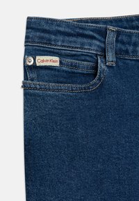 Vaqueros de denim azul oscuro de Calvin Klein, que presentan un bolsillo frontal, un botón de metal y una etiqueta con texto rojo. La tela tiene una textura de sarga clásica.