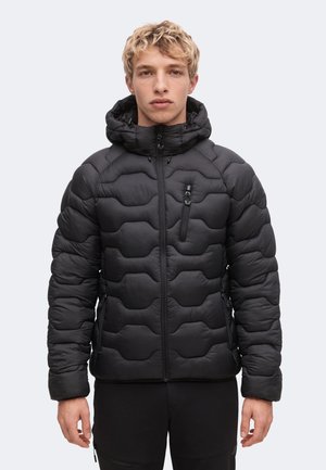 Giovane uomo che indossa una giacca imbottita nera con cappuccio e tasche frontali con zip, in piedi davanti a uno sfondo chiaro e uniforme.