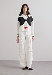 Un pull blanc avec un motif de nœud noir et des lèvres rouges, associé à un pantalon cargo large blanc comportant de multiples poches. Chaussures noires.