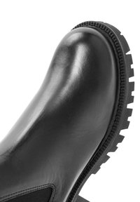 Bota de cuero negro con punta redondeada y suela de goma gruesa y estriada, que muestra un detallado cosido y panel lateral elástico.