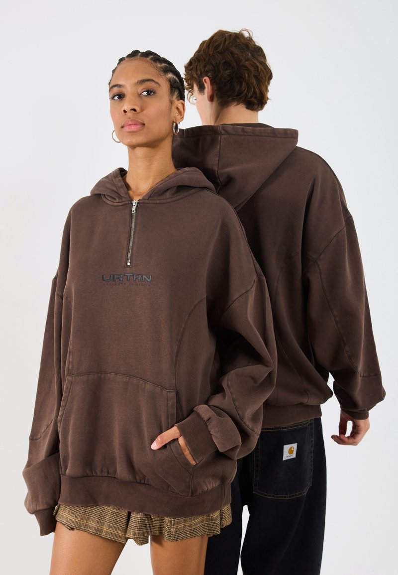 Sudadera con cremallera en color marrón, con capucha y bolsillo tipo canguro. Presenta puños y dobladillo de canalé. En la parte frontal se muestra el logo "URTRN". El modelo está de pie junto a una segunda persona.