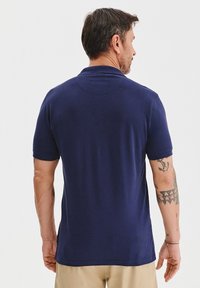 Granatowa koszulka polo z krótkimi rękawami, wykonana z gładkiego materiału bawełnianego. Posiada klasyczny kołnierzyk oraz dopasowany krój. Widoczne tatuaże na lewym ramieniu.